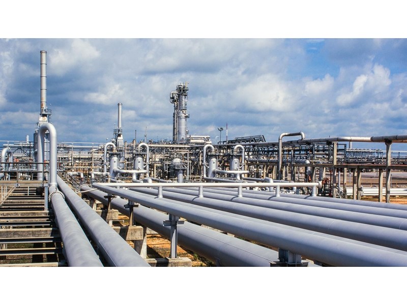 Close-up of a gas refinery, a network of pipelines and chimney stacks, against a cloudy blue sky.