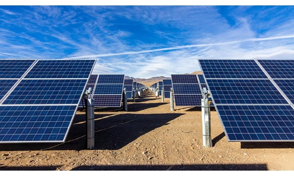 Rows of solar panels in the desert, with a mountainous backdrop and blue skies.