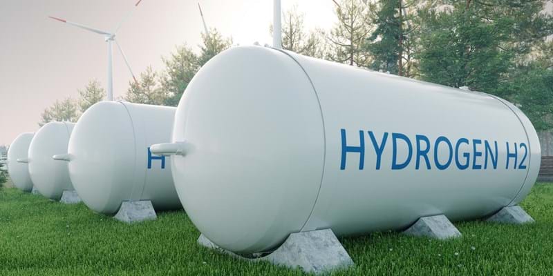 3D render of four hydrogen fuels tanks, in a tree-lined field with wind turbines to the side.