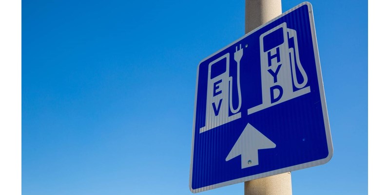 A blue and white road sign on a pole, displaying an area for EV and Hydrogen vehicle charging station.