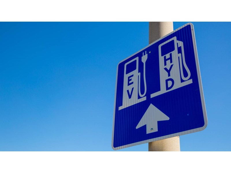 A blue and white road sign on a pole, displaying an area for EV and Hydrogen vehicle charging station.