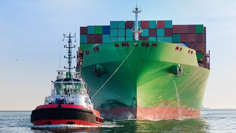 Front view on a tugboat towing a large cargo container ship with piled up cargo containers on deck into harbour.