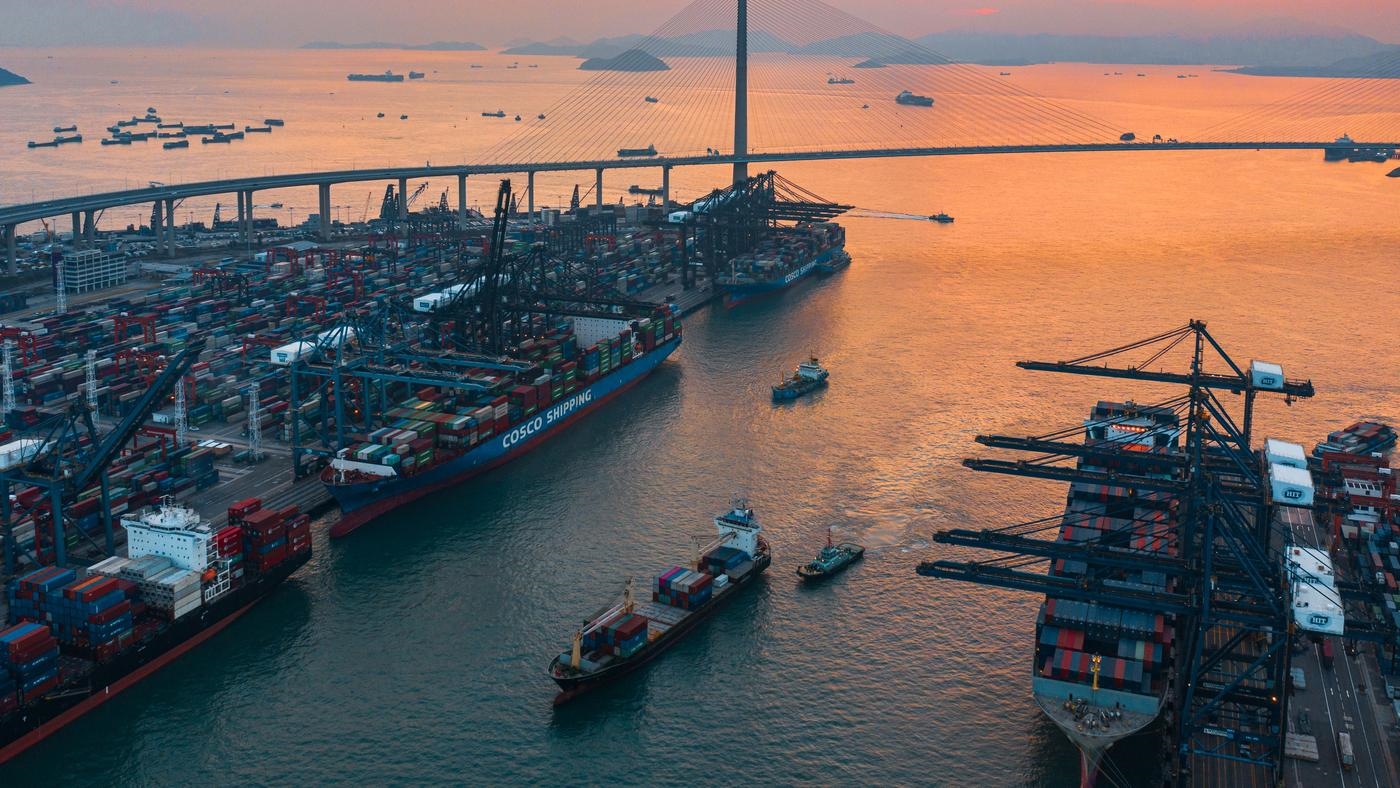A vast container port with large ships of cargo, next to a long modern suspension bridge.