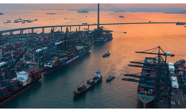 A vast container port with large ships of cargo, next to a long modern suspension bridge.