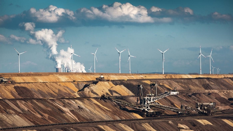Wind turbines sit atop a quarry, were machines are excavating, with smoke coming from the chimneys of a refinery in the background. 