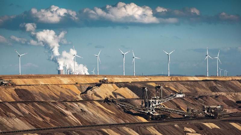 Wind turbines sit atop a quarry, were machines are excavating, with smoke coming from the chimneys of a refinery in the background.