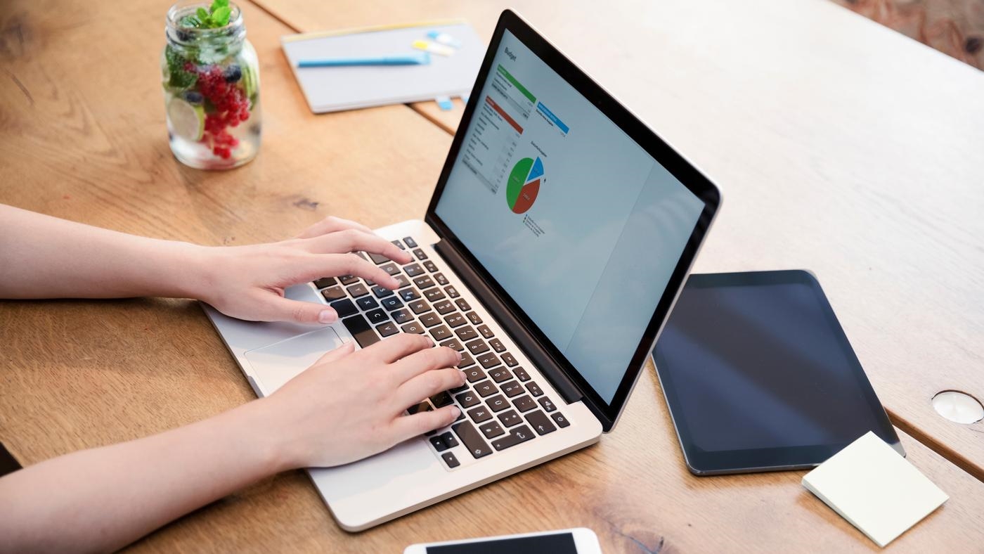 A person checking data on a laptop on a wooden table. iPad and smart phone on the desk.