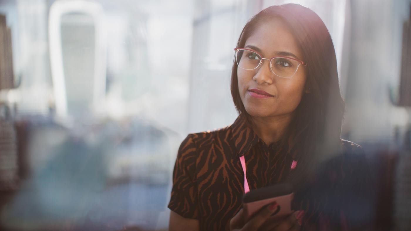 Thoughtful businesswoman with with black hair in eyeglasses using smart phone in sunny window, lens flare.