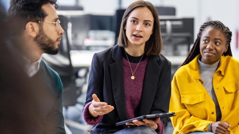 Young woman holding an iPad, sharing her ideas with team sitting in circle at office. Business team having meeting at work.