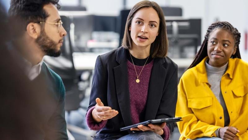 Young woman holding an iPad, sharing her ideas with team sitting in circle at office. Business team having meeting at work.