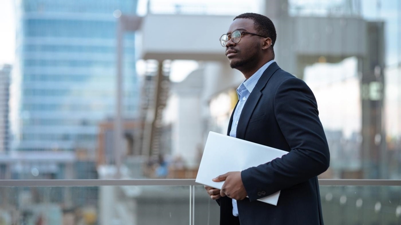 A young businessman on the balcony of the business building holding a laptop and thinking.