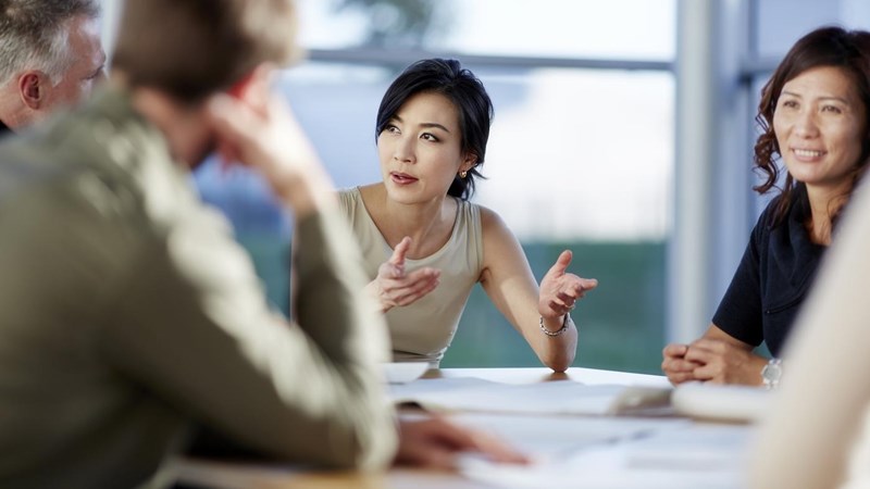 A group of people sitting around a table in a meeting, one lady is engrossed by the conversation.