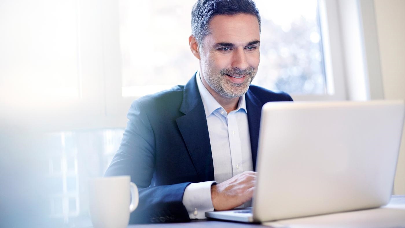 A smiling businessman working on a laptop, in front of window.