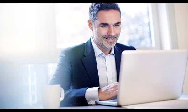 A smiling businessman working on a laptop, in front of window.