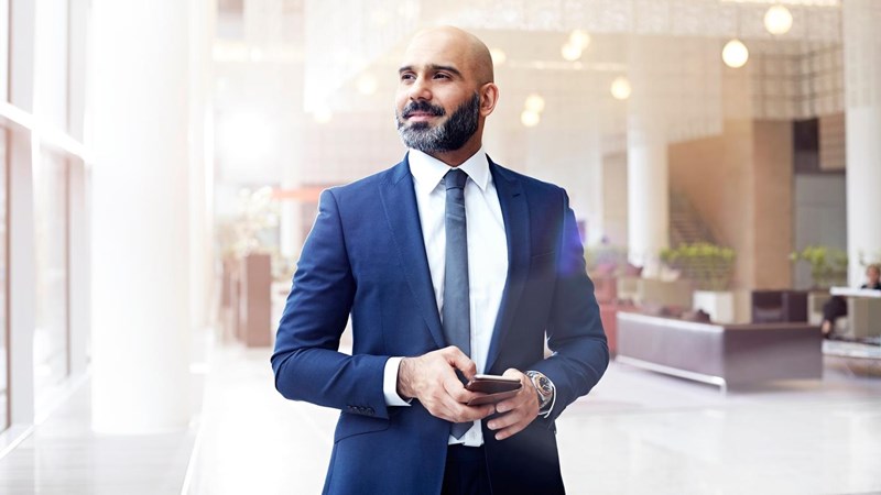 A businessman in a suit is holding a smartphone, in the lobby of an office building.