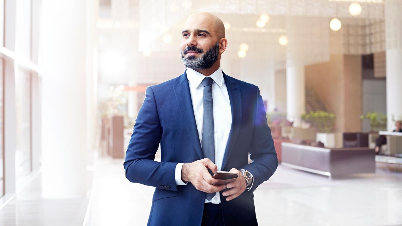 A businessman in a suit is holding a smartphone, in the lobby of an office building.