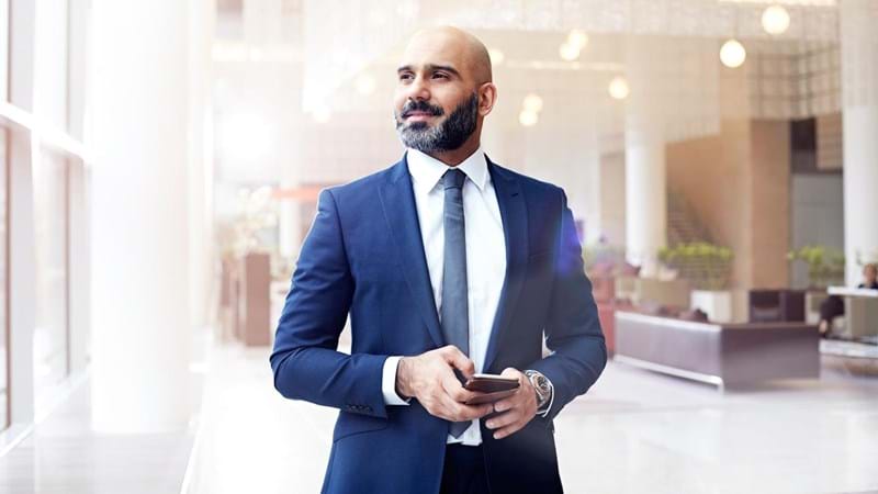 A businessman in a suit is holding a smartphone, in the lobby of an office building.