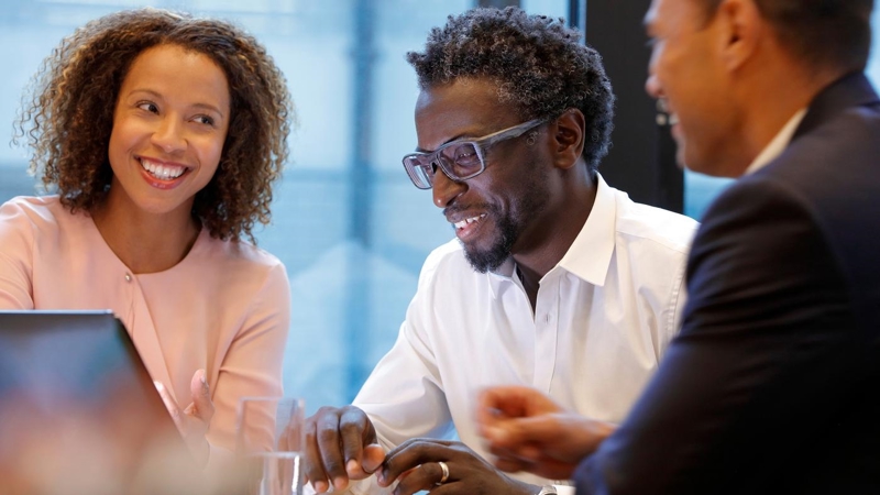 A group of business people smiling and sitting around a table looking at a laptop.
