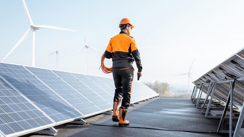 A worker standing on a roof next solar panels and wind turbines.