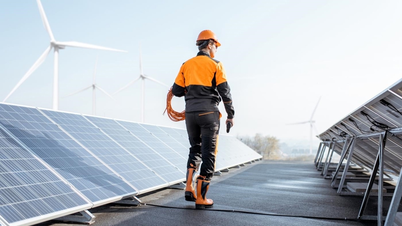 A worker standing on a roof next solar panels and wind turbines.