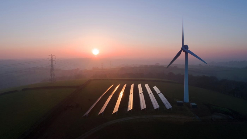 An aerial view of solar panels and a wind turbine in a field at sunset.