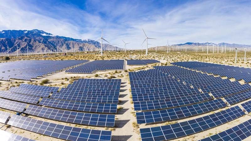 Solar panels and wind turbines in the desert, surrounded by a mountain range, against the blue skies.