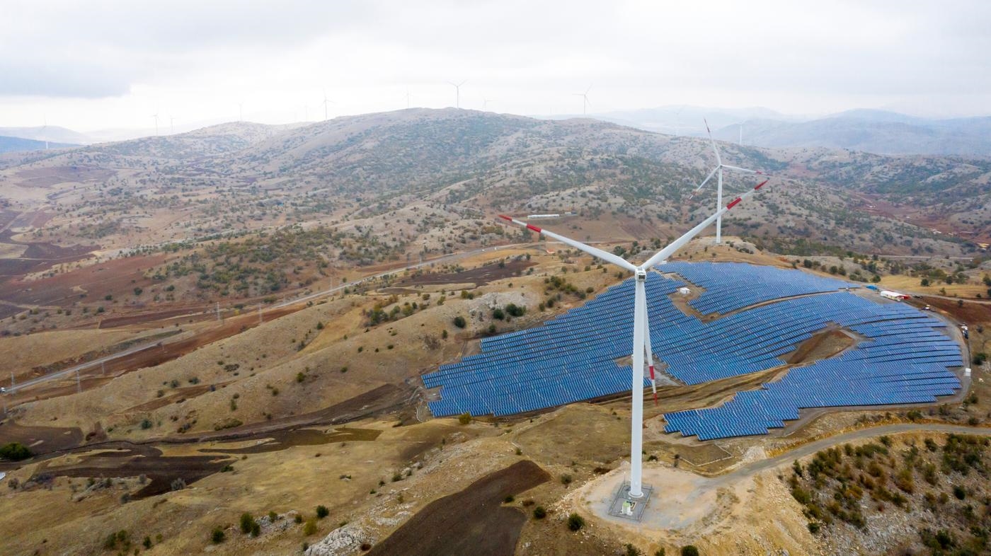 An aerial view of wind turbines and solar panels, across rolling hills.