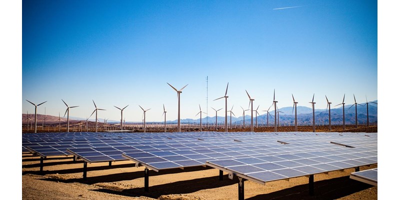 Solar panels and wind turbines in the desert on a backdrop of mountains and clear blue skies.