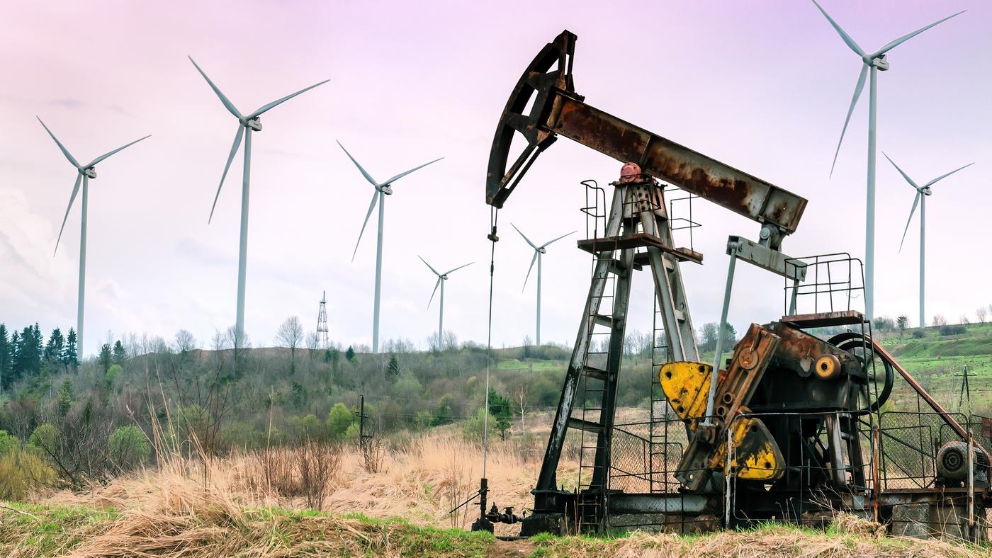 An oil pump in overgrown fields with wind turbines in the background.