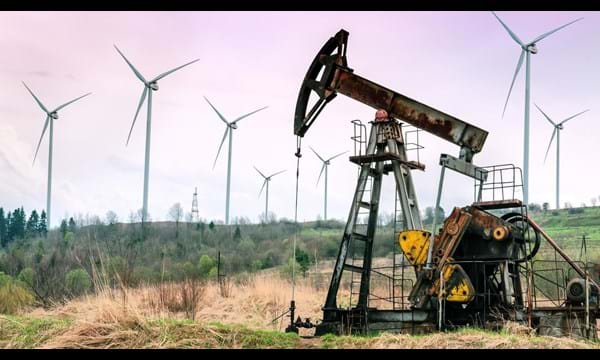 An oil pump in overgrown fields with wind turbines in the background.