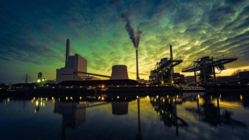 A energy refinery is reflected in a body of water, against an ominous looking green sky.
