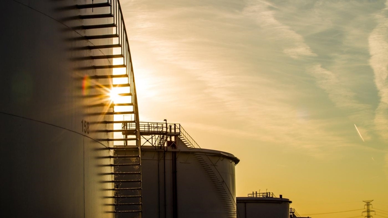 The sun is setting behind a row of oil tanks.