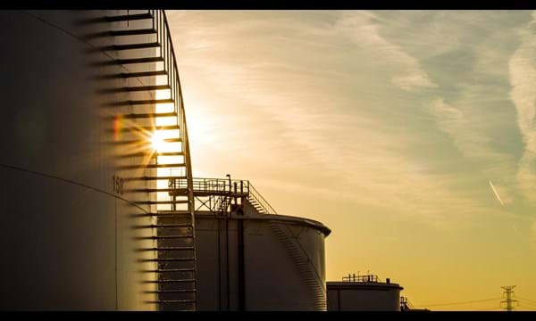The sun is setting behind a row of oil tanks.
