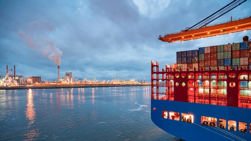 A large container ship docked at a port, next to a large oil refinery.