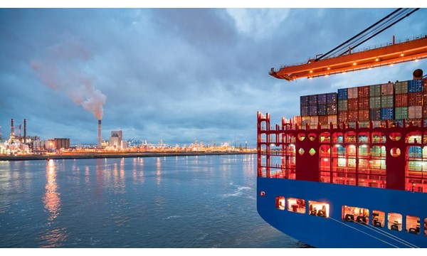 A large container ship docked at a port, next to a large oil refinery.