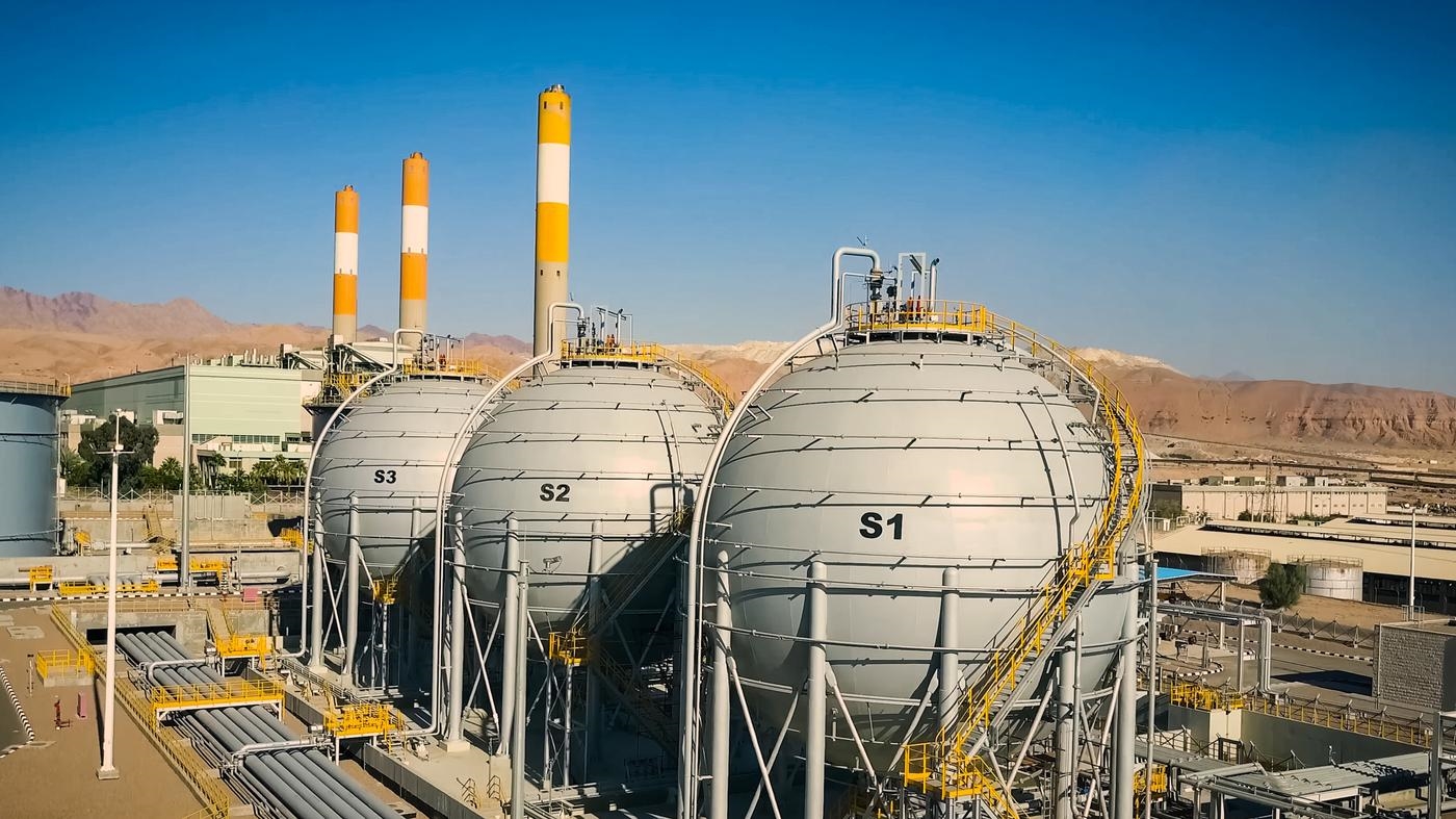 A large power plant with three LNG tanks, mountain range and blue skies in the background.