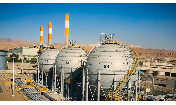 A large power plant with three LNG tanks, mountain range and blue skies in the background.