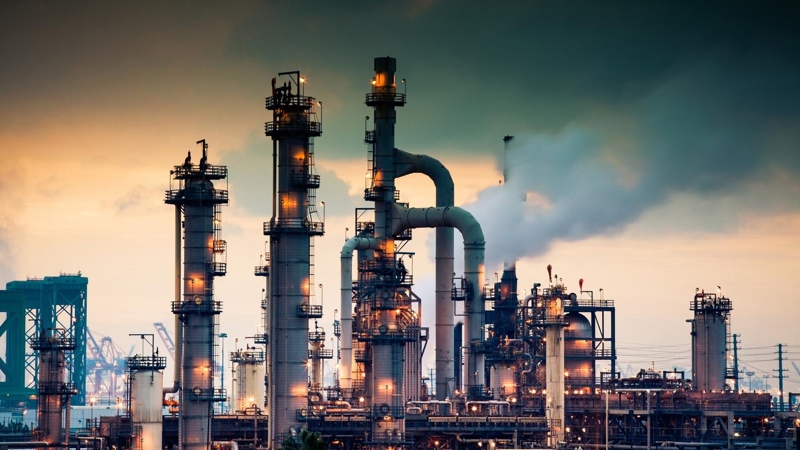 A steaming, glowing mass of smoke stacks and distillation towers connected by conduit and pipe racks towers in the early morning light in front of the Port of Los Angeles.