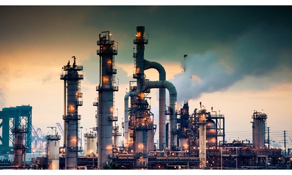A steaming, glowing mass of smoke stacks and distillation towers connected by conduit and pipe racks towers in the early morning light in front of the Port of Los Angeles.