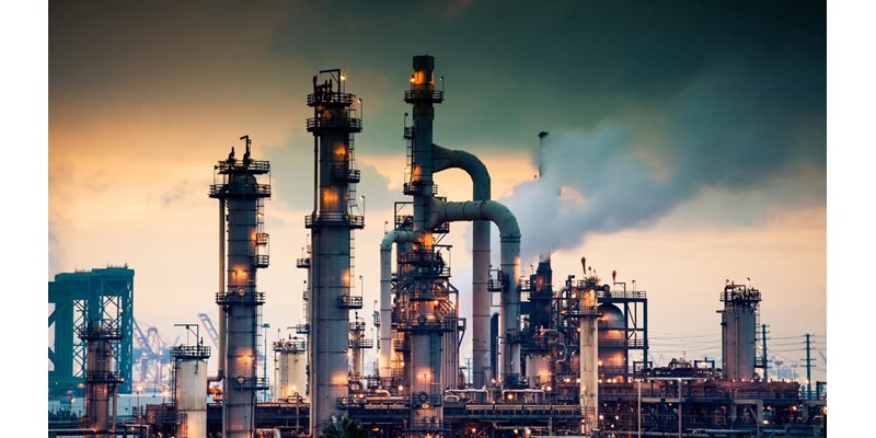 A steaming, glowing mass of smoke stacks and distillation towers connected by conduit and pipe racks towers in the early morning light in front of the Port of Los Angeles.