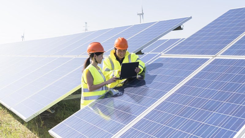 Two engineers in hard hats looking at a laptop on a section of solar panels, wind turbines and electricity pylons in the background.