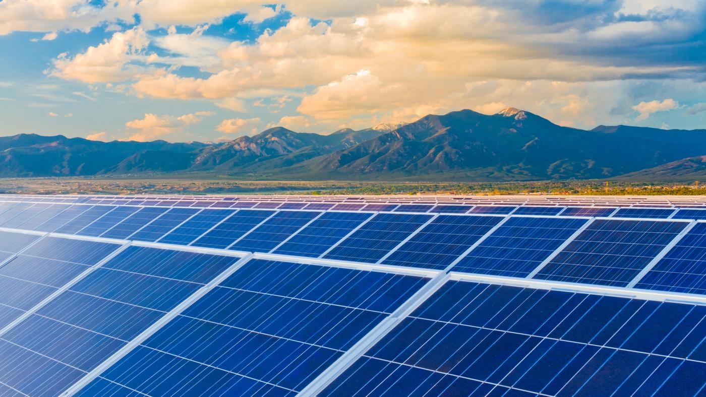 Rows of solar panels in a field, with mountains and blue skies in the background.