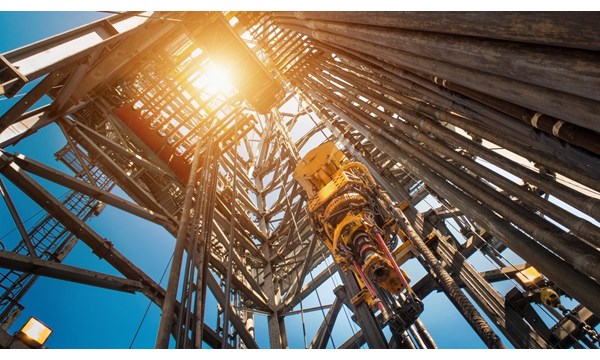 An upward view of a central drilling column of an oil rig, the sun and blue skies in the background.