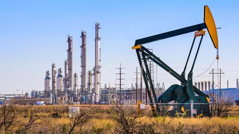 An oil pump in front of an oil refinery, with a network of electricity pylons.