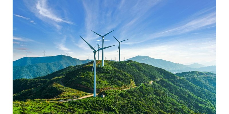 Wind turbines on top of tree covered mountains, against blue skies.