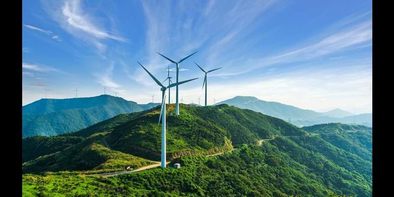 Wind turbines on top of tree covered mountains, against blue skies.