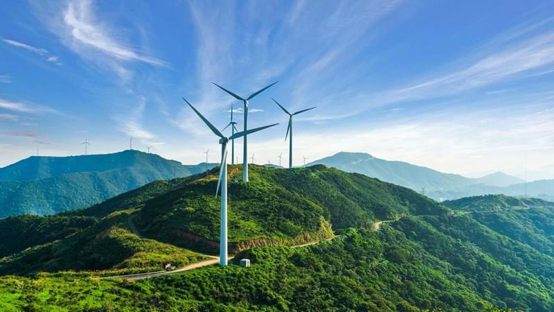 Wind turbines on top of tree covered mountains, against blue skies.